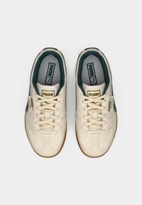White leather sneakers with a textured finish, green accents, and a rubber sole. Features gold logo and lace closure. Slim, rounded shape.