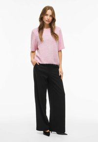 Gaiši rozā, īss piedurkņu adīts džemperis ar mīkstu tekstūru, kombinēts ar melnām plašu kāju biksēm un melniem smailiem apaviem.