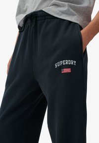Pantalones deportivos negros de mezcla de algodón, con cintura elástica, bolsillos laterales y logo bordado de "SUPERDRY" con un pequeño detalle de bandera.