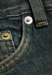 Gros plan sur un tissu en denim bleu avec des coutures jaunes et deux rivets métalliques sur le bord d'une poche et une boucle de ceinture.