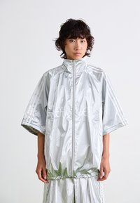 Zilveren, lichte jacket met korte mouwen, hoge kraag en rits. Heeft drie witte strepen op de mouwen en een logo met tekstpaneel.