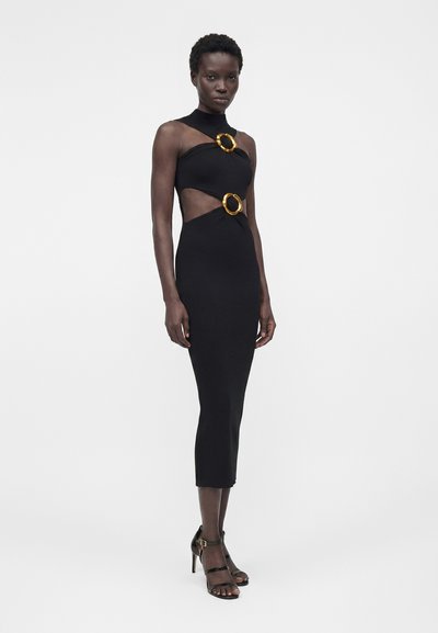 Γυναίκα που φοράει εφαρμοστό μαύρο midi φόρεμα με κοψίματα και δύο μεγάλες χρυσές κυκλικές αγκράφες, συνδυασμένο με μαύρα σανδάλια με ψηλά τακούνια.