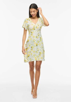 Kvinde iført en hvid, kortærmet kjole med V-udskæring og gul citronprint, gående med den ene hånd, der rører ved hendes hår, og gennemsigtige hæle.