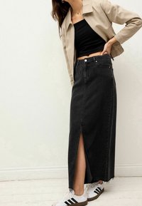 Jupe maxi en denim noir avec une fente à l'avant, un ourlet effiloché et cinq poches, portée avec un crop top noir et des baskets blanches avec des rayures noires.