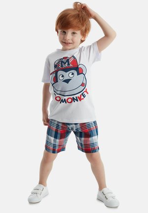 Weißes T-Shirt mit einem Affenmotiv und Text, kombiniert mit karierten Shorts in Rot, Blau und Weiß; abgerundet mit weißen Slippern.