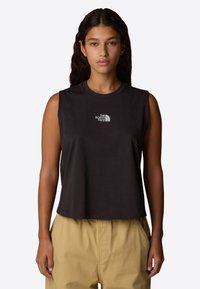 Zwart mouwloos crop top van katoen, met een ronde hals en een wit logo op de borst. Gekleed met beige broek.