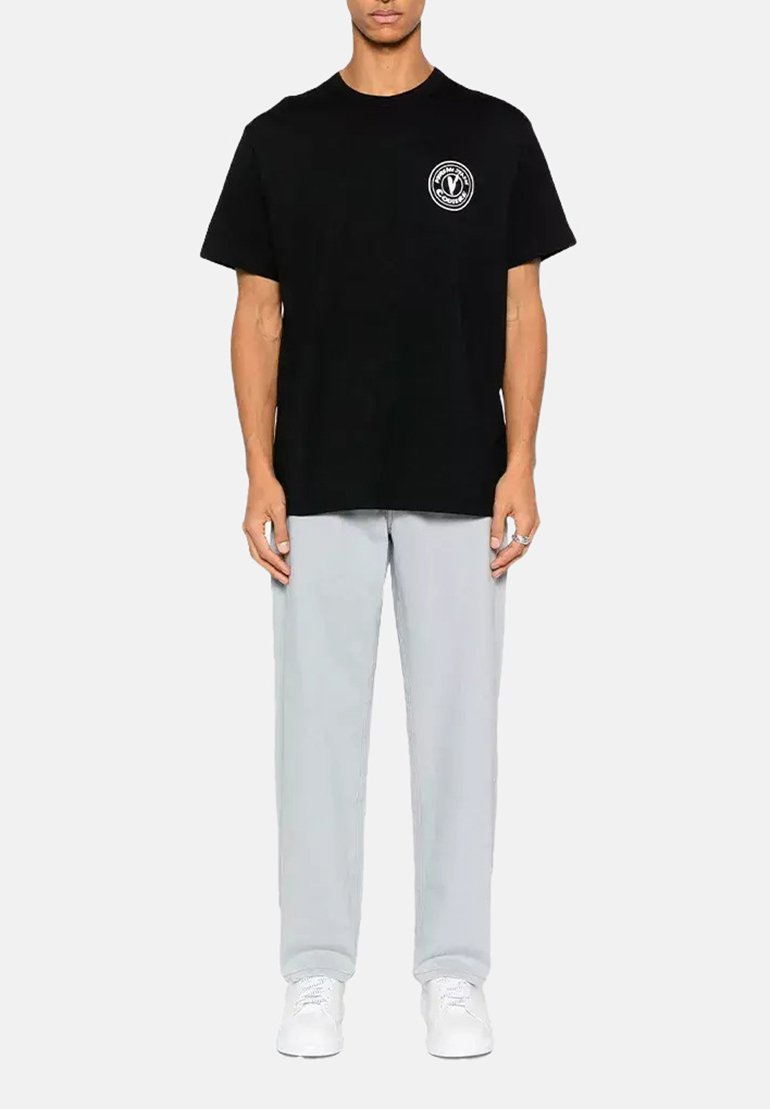 T-shirt nera in cotone con un logo rotondo sul petto, abbinata a pantaloni grigi chiari. Il modello indossa sneaker bianche ed è posizionato davanti a uno sfondo neutro.