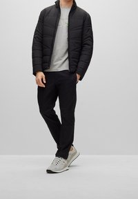 Svart puffjacka med kviltat mönster, grå t-shirt med grönt logotyp, svarta byxor och ljusgrå sneakers med nät och solida detaljer.