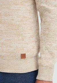 Beige strikkegenser med ribbestrikket kant og mansjetter, med en liten brun lærmerke med "Blend"-logo, brukt sammen med mørkeblå jeans.
