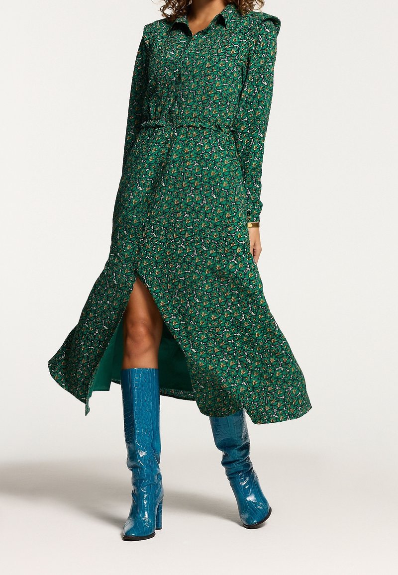 Robe longue à manches longues à fleurs vertes avec un devant à boutons, une taille ceinturée et une fente sur le côté. Associée à des bottes brillantes bleu à hauteur de genou.