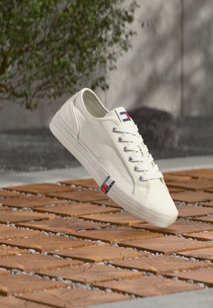 Baskets en toile blanche avec un bout arrondi, comprenant des œillets métalliques, des lacets blancs et un petit logo bleu, rouge et blanc. Semelle en caoutchouc texturée.