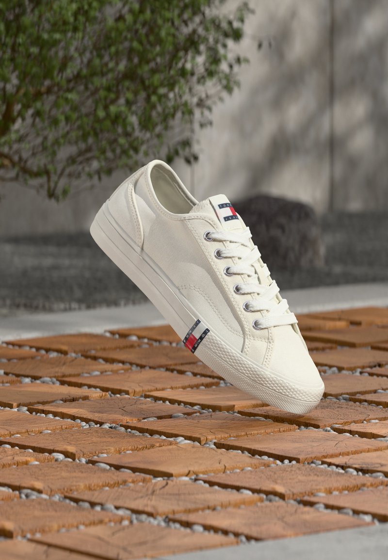 Baskets en toile blanche avec un bout arrondi, comprenant des œillets métalliques, des lacets blancs et un petit logo bleu, rouge et blanc. Semelle en caoutchouc texturée.