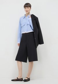 Camicia corta a righe blu chiaro, pantaloni larghi neri, blazer nero appoggiato sulla spalla e ballerine nere con una fascia.