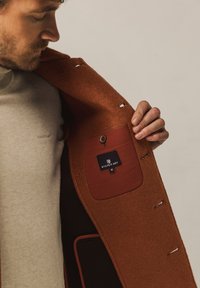 Bruine wollen jas met een wollen textuur, voorzien van een knoopzakje aan de binnenkant en een label. Model draagt een lichtgrijze trui eronder.
