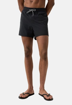 Mand iført sorte badebukser med snøre og sorte klipklappere, stående foran en ensartet hvid baggrund.