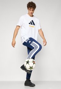 Hvid t-shirt med sort og gul "REAL MADRID" logo, parret med marinblå bukser med hvide striber og sorte sportssko, som jonglerer med en fodbold.
