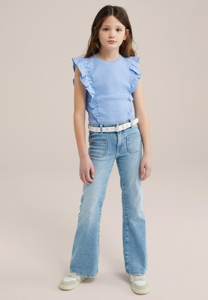 Hellblaues, rüschengeschnittenes Oberteil mit kurzen Ärmeln, kombiniert mit ausgestellten Jeans. Das Outfit wird durch einen weißen Gürtel und hellgrüne Sneakers ergänzt.