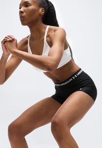 Αθλητική γυναίκα με λευκό αθλητικό σουτιέν και μαύρο σορτς Under Armour εκτελεί καθίσματα μπροστά σε απλό ανοιχτό φόντο.