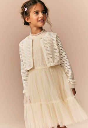Junges Mädchen mit geflochtenen Haaren, das ein cremefarbenes Spitzenkleid und eine passende gestrickte kurze Strickjacke mit einer kleinen Blumen-Haarspange trägt.