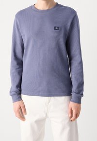 Man som bär en långärmad, texturerad lila skjorta med en liten Calvin Klein-logotyp på bröstet och vita byxor.