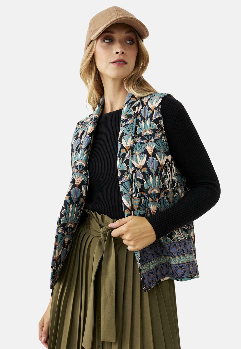 Mujer con gorra beige, blusa de manga larga de canalé negra, chaleco con estampado floral y falda larga de cintura alta plisada en verde oliva con cinturón de lazo.