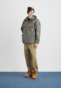 Jachetă anorak de camuflaj în nuanțe de verde și maro, asortată cu pantaloni de lucru bej și cizme maro, având un buzunar frontal și glugă.