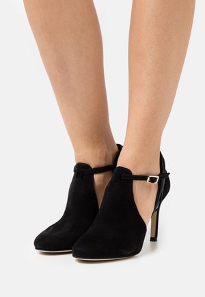 Černé nubukové kotníkové boty s designem T-řemínku, špičatou špičkou a stiletto podpatkem. Zlatá spona na řemínku. Hladká textura.