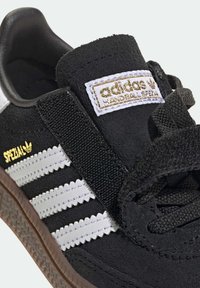 Black Adidas sneaker featuring a suede upper, white leather stripes, a textured black strap, and a brown rubber sole. Gold logo accent.
