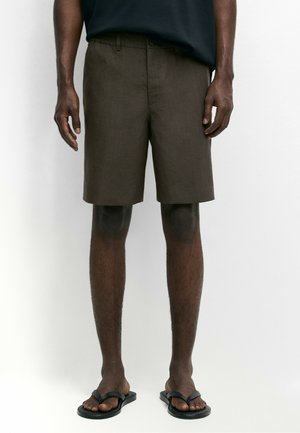 Person trägt braune knielange Shorts, schwarze Flip-Flops und ein dunkles Oberteil und steht vor einem einfarbigen weißen Hintergrund.
