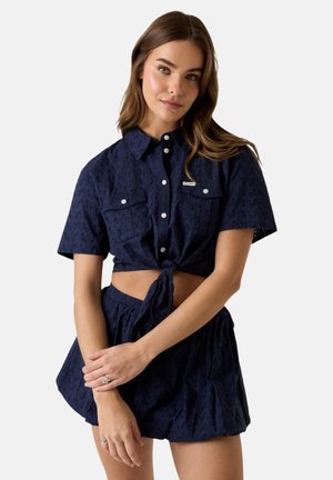 Donna con capelli castani lunghi che indossa un top corto blu navy a maniche corte con bottoni, annodato in vita, e shorts abbinati, in piedi con le braccia incrociate.