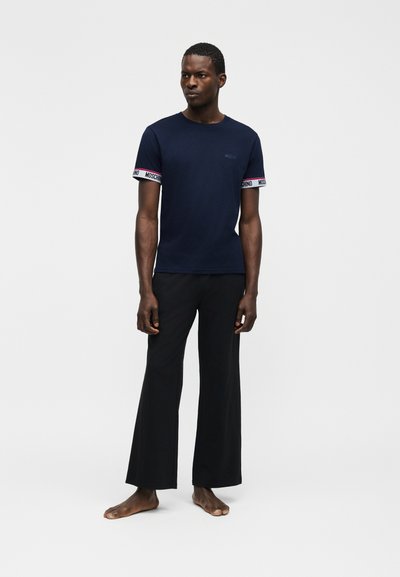 Άνδρας που στέκεται ξυπόλυτος, φορώντας μπλε ναυτικό t-shirt με επωνυμικά μανσέτες και φαρδιά μαύρη παντελόνι, μπροστά σε απλό λευκό φόντο.