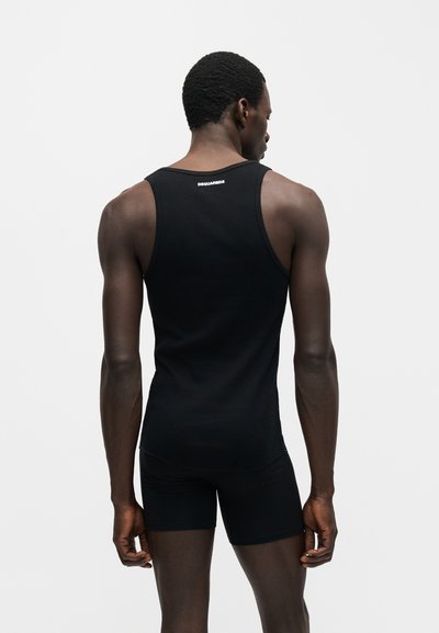 Άνδρας με μαύρο αμάνικο μπλουζάκι και σορτς στέκεται με την πλάτη στην κάμερα σε μονόχρωμο φωτεινό φόντο.