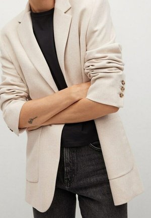 Person, die einen beigen Blazer mit hochgekrempelten Ärmeln über einem schwarzen Hemd und dunklen Jeans trägt, Arme verschränkt, zeigt ein kleines Rosen-Tattoo am linken Unterarm.