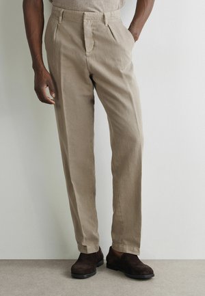 Mann trägt beige plissierte Hose und dunkelbraune Wildlederschuhe, steht mit einer Hand in der Tasche vor hellem Hintergrund.