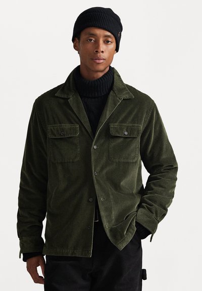 Jachetă din corduroy de culoare verde măslin, cu două buzunare pe piept, design cu guler și accesorii din metal argintiu. Purtați peste un pulover negru cu dungi.