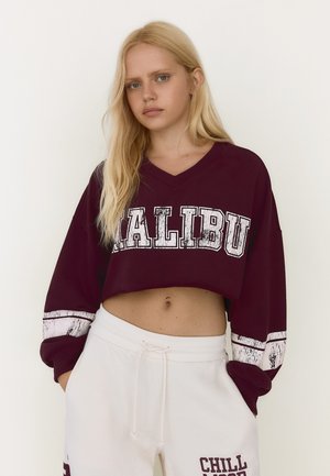 Ung kvinde med langt blondt hår iført en vinrød cropped sweatshirt med "MALIBU" og hvide joggingbukser med "CHILL MODE" tekst.