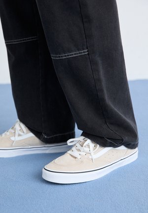 Personne portant des baskets beige et blanches avec des lacets blancs et un pantalon large noir debout sur un sol bleu clair.