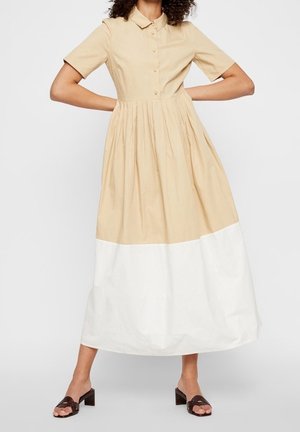 Sukienka maxi z guzikiem z przodu. Beżowa góra, biała spódnica. Krótkie rękawy i marszczona talia. Noszona z brązowymi sandałami na obcasie.