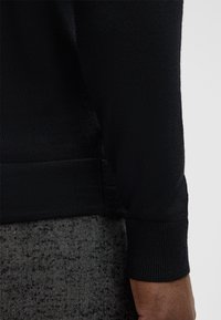 Schwarzer Strickpullover mit strukturierter Oberfläche und gerippten Bündchen, getragen über einem grau karierten Kleidungsstück mit Fischgrätenmuster, wobei nur ein Teil des Ärmels sichtbar ist.