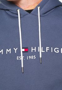 Felpa blu con lacci bianchi che presenta il logo "Tommy Hilfiger" e una bandiera rossa, bianca e blu sopra il testo "EST. 1985" sul petto.