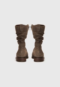 Bottes en daim marron arrivant mi-mollet avec un design flou. Comprend une fermeture éclair sur le côté, un talon en bois plat et un revers roulé pour apporter texture et détail.