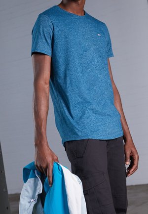 Mężczyzna w niebieskim melanżowym T-shircie i czarnych spodniach cargo, trzyma niebiesko-białą kurtkę w lewej ręce na szarym tle.
