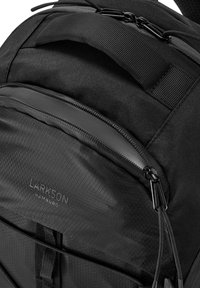 Schwarzer Rucksack mit strukturiertem, sechseckigem Muster, ausgestattet mit einer vorderen Reißverschlusstasche und verstellbaren Trägern. Logo auf der Vorderseite angebracht.