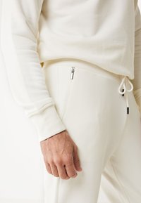 Vit bomullströja med rundad hals och ribbade muddarmar, kombinerad med matchande joggers som har dragkedjefickor och dragsko i midjan.