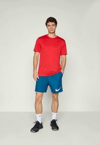 T-shirt de sport rouge avec logo noir, short bleu avec logo blanc, chaussures noires et chaussettes blanches. Design simple, tissu léger.