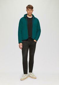 Teal softshell jack met een capuchon, rits aan de voorkant en twee zakken. Zwarte lange mouw shirt eronder, zwarte slim-fit jeans en witte sneakers.