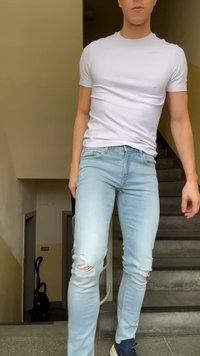 Νεαρό άτομο που φοράει εφαρμοστή λευκή μπλούζα, ανοιχτόχρωμο σκισμένο τζιν και σκούρα αθλητικά παπούτσια στέκεται σε εσωτερική σκάλα.