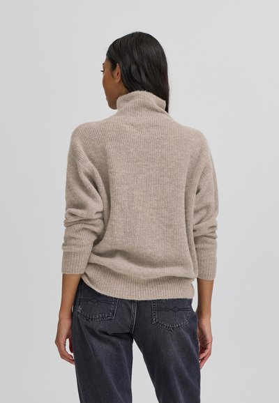 Béžový žebrovaný svetr s vysokým límcem a uvolněným střihem, spárovaný s tmavě šedými džínami s prošívanými detaily na zadních kapsách.