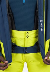 Waterdichte skijas met een marineblauwe buitenkant en heldergele broek. Beschikt over drukknoopsluitingen, een verstelbare tailleband en een ritssluiting aan de voorkant.