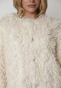 Biały sweter z kręconym splotem o luźnym kroju, z dużymi guzikami i okrągłym dekoltem. Materiał wydaje się miękki i puszysty.